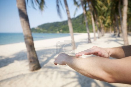 Photo for Close-up of hand while applying sunscreen. Skin care during summer vacations on beautiful sand beach with palm trees in tropical destination - Royalty Free Image