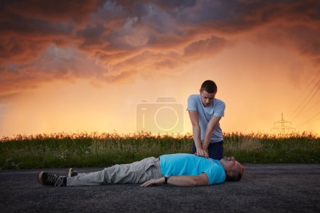 Photo for Dramatic resuscitation on rural road. Young man provides first aid to unconscious middle aged man under storm clouds. Themes rescue, help and hope - Royalty Free Image