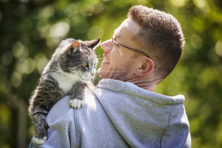 Foto de Sonriente hombre cargando a su curioso gato en el hombro. Propietario de mascotas con gato viejo tabby en el jardín en el día soleado - Imagen libre de derechos