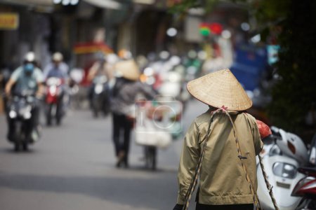 Téléchargez les photos : Accent sélectif sur le chapeau conique traditionnel de la personne marchant contre les motos de la circulation dans la rue animée dans le vieux quartier de Hanoi, Vietnam - en image libre de droit