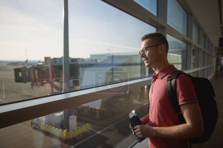 Foto de Un hombre sonriente viajando en avión. Pasajero mirando a través de la ventana en la pista del aeropuerto mientras sostiene la billetera con tarjeta de embarque en su mano - Imagen libre de derechos