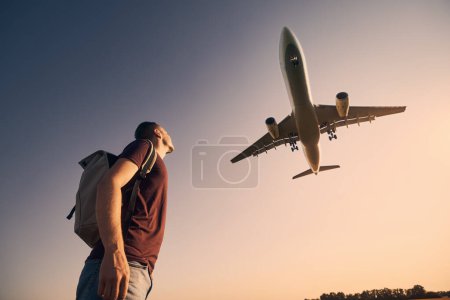 Foto de Hombre con mochila mirando hacia arriba para aterrizar en el aeropuerto durante el hermoso atardecer de verano. - Imagen libre de derechos