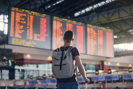 Photo for Traveling by airplane. Man walking with backpack and suitcase walking through airport terminal and looking at departure information - Royalty Free Image