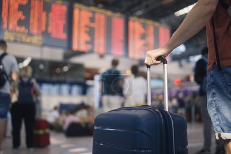 Photo for Traveling by airplane. Man waiting in airport terminal. Selective focus on hand holding suitcase against arrival and departure board. Passenger is ready for travel - Royalty Free Image