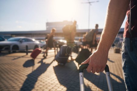 Gruppe von Menschen zu Fuß zum Flughafen-Terminal bei Sonnenuntergang. Selektiver Fokus auf die Hand des Mannes mit Koffer.