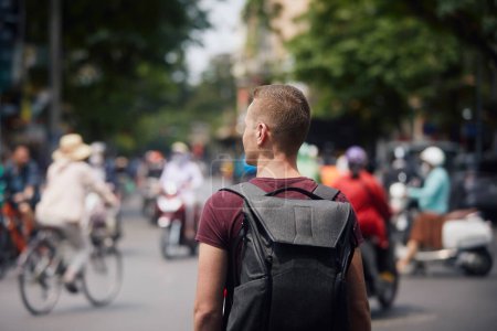 Foto de Viajero con mochila descubriendo Vietnam. Hombre en medio de una concurrida calle llena de motos y bicicletas en Hanoi, Vietnam - Imagen libre de derechos