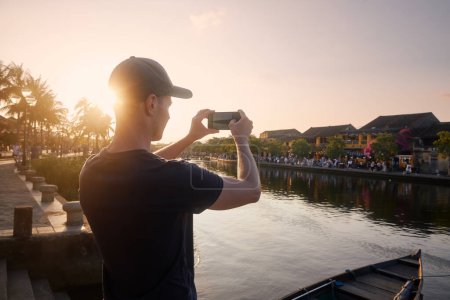 Photo for Man photographing Hoi An cityscape. Tourist with smart phone in ancient city during beautiful sunset, Vietnam - Royalty Free Image