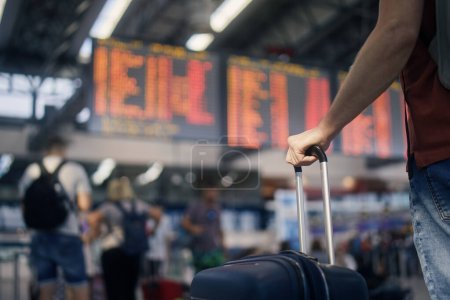 Photo for Traveling by airplane. Man waiting in airport terminal. Selective focus on hand holding suitcase against arrival and departure board. Passenger is ready for travel. - Royalty Free Image