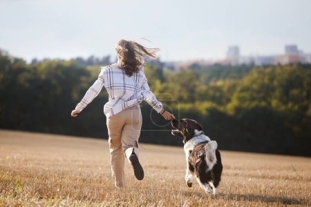 Photo for Rear view of happy teen girl running with her dog across field against cityscape. Joyful pet owner with Czech Mountain Dog - Royalty Free Image
