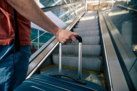 Photo for Traveling by airplane. Man standing on escalator in airport terminal. Selective focus on hand holding suitcase. Passenger is ready for travel. - Royalty Free Image