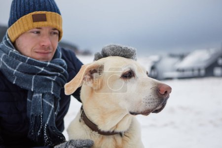 Téléchargez les photos : Homme vêtu d'une veste chaude, avec écharpe et casquette caressant son chien. Propriétaire d'animal de compagnie et labrador récupérateur contre village de montagne en hiver. Focus sélectif sur le chien - en image libre de droit