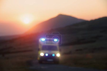 Téléchargez les photos : Vue déconcentrée de la voiture d'ambulance du service médical d'urgence sur la route de montagne au lever du soleil. Thèmes santé, secours et urgence - en image libre de droit
