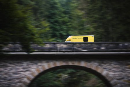 Photo for Ambulance car of emergency medical service in blurred motion on stone bridge in mountains. Themes rescue, urgency and  health care - Royalty Free Image