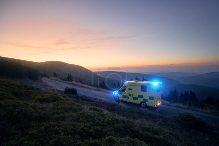Foto de Ambulancia coche del servicio médico de emergencia carretera de montaña contra el amanecer. Cielo mohoso con espacio para copiar. Temas de rescate, urgencia y atención sanitaria - Imagen libre de derechos