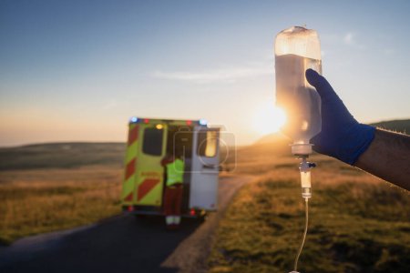 Foto de Primer plano de la mano de paramédico sosteniendo goteo IV contra coche ambulancia del servicio médico de emergencia. Temas de rescate, urgencia y atención sanitaria. - Imagen libre de derechos