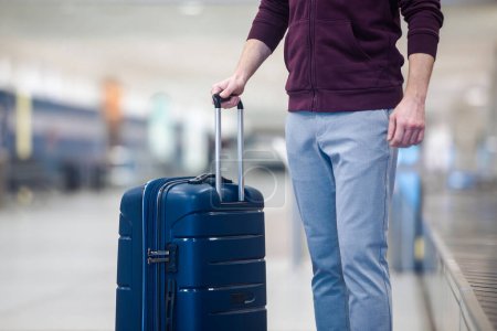 Téléchargez les photos : Passager voyageant en avion ramassant la valise bleue de la bande transporteuse dans le terminal des arrivées de l'aéroport - en image libre de droit