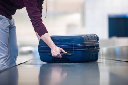 Téléchargez les photos : Passager voyageant en avion ramassant la valise bleue de la bande transporteuse dans le terminal des arrivées de l'aéroport - en image libre de droit
