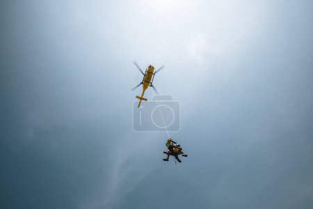 Téléchargez les photos : Deux ambulanciers suspendus à une corde sous le service médical d'urgence d'hélicoptère volant. Thèmes sauvetage, aide et héros. Ciel avec espace de copie - en image libre de droit