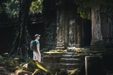 Foto de Viajero en Camboya. Hombre con mochila admirando las ruinas del antiguo templo cubierto de vegetación tropical. - Imagen libre de derechos