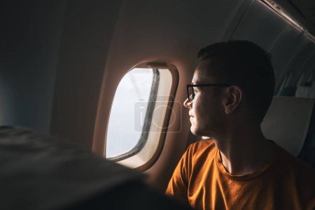 Téléchargez les photos : Portrait d'un homme voyageant en avion. Passager regardant par la fenêtre de l'avion pendant le vol - en image libre de droit
