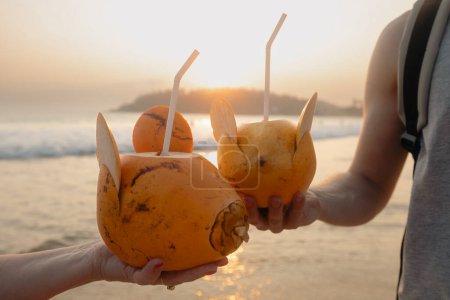 Man and woman drinking fresh coconut water together on idyllic sand beach. Healthy natural refreshment at sunset. Mirissa in Sri Lanka