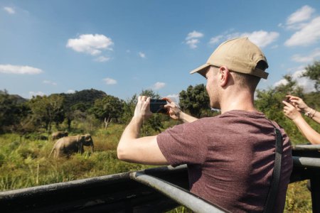 Téléchargez les photos : Tourist photographie un troupeau d'éléphants sauvages avec un téléphone intelligent. Homme profitant d'un safari au Sri Lanka - en image libre de droit