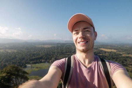 Photo for Happy man taking selfie photo from summer vacation day. Handsome tourist wearing cap and smiling at camera against tropical landscape. - Royalty Free Image
