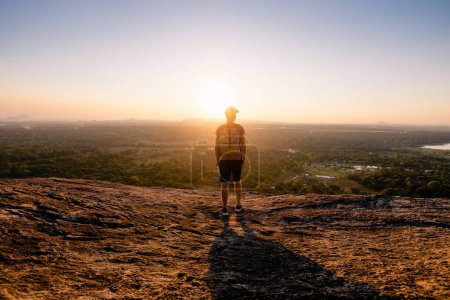 Foto de Vista trasera del hombre con mochila en roca con vistas a la puesta del sol sobre el paisaje tropical. Viajar a Sri Lanka - Imagen libre de derechos