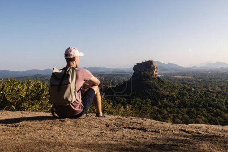 Téléchargez les photos : Homme avec sac à dos assis sur le rocher et regardant le paysage. Beau paysage avec Sigiriya rock. Voyageur solo en Sri Lanka. - en image libre de droit