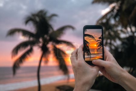 Foto de Primer plano de las manos sosteniendo el teléfono inteligente. Hombre fotografiando puesta de sol sobre el océano con teléfono móvil. Palmeras de coco en la playa de arena en la costa sur de Sri Lanka - Imagen libre de derechos
