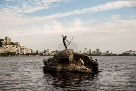 Foto de Río de Janeiro, Brasil - 10 de septiembre de 2023: Estatua de pescador indígena señalando su arco y flecha hacia el agua de la laguna Rodrigo de Freitas con el distrito de Ipanema en el horizonte. - Imagen libre de derechos