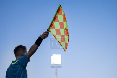 Photo for Raised flag of the sideline referee during football match, blue sky and lamps in the background. - Royalty Free Image