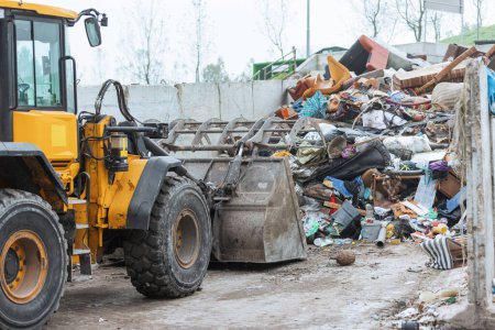 Photo for Yellow wheel loader, with lifted scrap grapple, moving along the recycling center area in process handling dumped waste - Royalty Free Image