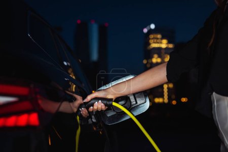 Photo for Female hands opening an electric car charging socket cap and plugging in a charger, visible skyscrapers shining in a summer night in the background, close up shot. - Royalty Free Image