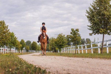 Reiterin, Reiterin auf dem Weg, der zwischen Zäunen aus weißem Holz und Feldern führt. Freizeitkonzept für den Pferdesport.