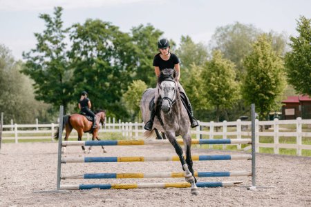 Photo for Girl rider on dapple gray horse jumping over triple bars in outdoor arena on a sunny summer day. Horse sports competition concept. - Royalty Free Image