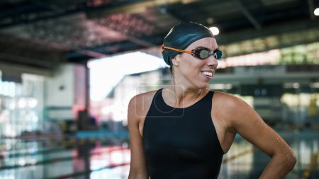 Professional female swimmer in a black swimsuit, cap, and goggles, looking around, smiling, and focusing before the race start.