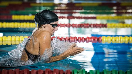 Atleta femenina nadando en estilo de pecho en el carril de la piscina, acariciando, sumergiendo y levantando del agua para respirar.