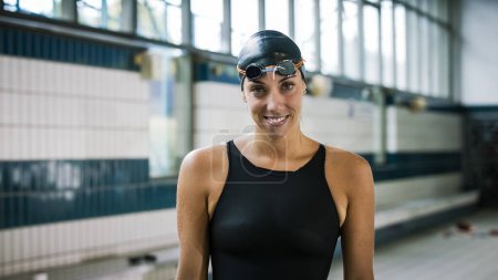 Professional female swimmer in a black swimsuit, cap, and goggles, looking around, smiling, and focusing before the race start.