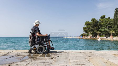Tourist man with a disability sitting on the dock and looking at the open sea. Wheelchair travel and accessible tourism concept.