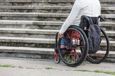 Mujer en silla de ruedas que se detiene y espera en la parte inferior de la escalera de calle inaccesible, incapaz de subir. Conceptos de dificultad y discapacidad.