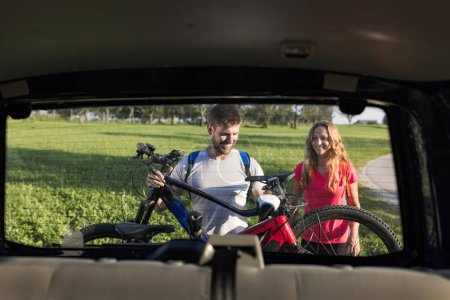 Inside car view of a young couple, the electric mountain bikers, a man unloading EMTB from the hitch rack on the rear of the vehicle.