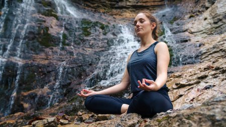 Calm girl practicing yoga, sitting in sukhasana pose while beautiful waterfall burbling in the background. Serenity, nature, and healthy lifestyles concepts.