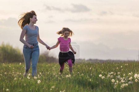 Mère et sa petite fille courant la main dans la belle prairie verte au coucher du soleil.
