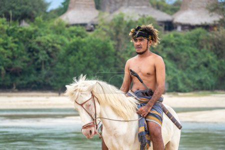 Téléchargez les photos : Un membre de la tribu Marapu Sumba monte gracieusement un cheval le long du rivage, avec la captivante cabane traditionnelle Ratenggaro en toile de fond - en image libre de droit