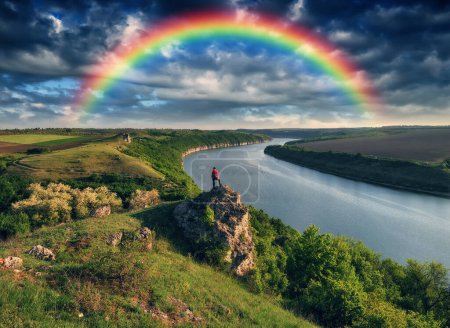 Tourist standing on the edge of the cliff and looking at the rainbow. Rainbow over the river. Nature of Ukraine