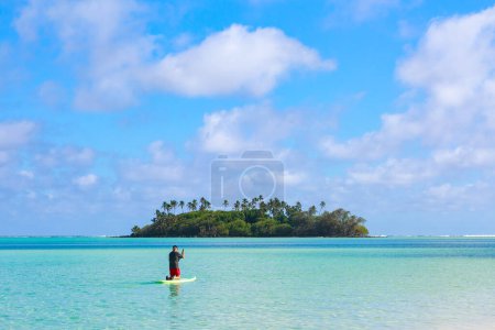Photo for A man paddleboards towards a tropical island in the middle of a beautiful turquoise lagoon. Photographed at Muri, Rarotonga, Cook Islands, looking towards Taakoka Island - Royalty Free Image