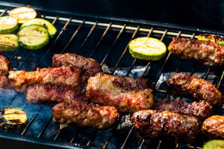 Téléchargez les photos : Griller les rouleaux de viande appelés mici ou mititei avec des légumes sur le barbecue de l'omble. Grill au charbon avec feu brûlant - en image libre de droit