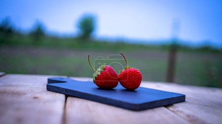 Nahaufnahme von zwei Erdbeeren auf einem kleinen schwarzen Schneidebrett isoliert im Freien auf einem Holztisch.