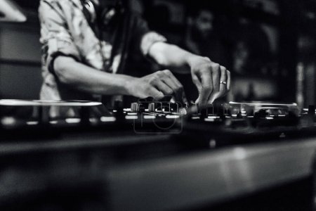 DJ is playing music in a nightclub. You can see his hands mixing music with his dj mixer and color lights os a party in the background.